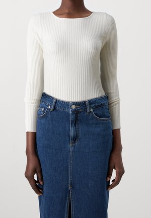 Persoon die een wit geribd top met lange mouwen draagt, ingestopt in een hooggetailleerde blauwe denim rok met een voorste split en knoopsluiting.