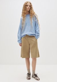 Hellblauer Strickpullover mit gerippten Bündchen, beigefarbene knielange Shorts, graue Socken und gemusterte Schuhe in Braun und Weiß.