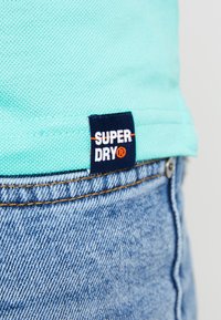 T-shirt en coton turquoise clair avec un ourlet et une petite étiquette marine marquée « SUPERDRY » avec des accents rouges ; associé à un jean en denim bleu clair.