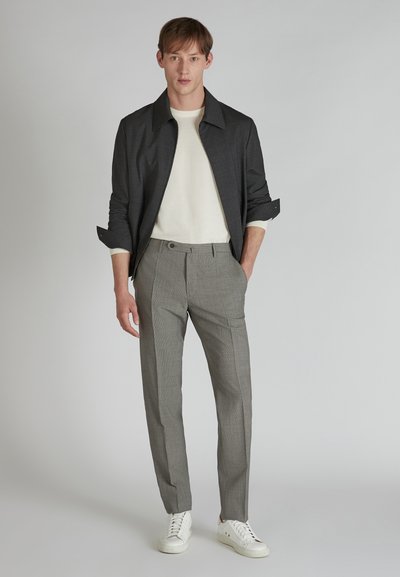 Pantalons gris à carreaux, chemise à col rond blanche et veste sombre. Le modèle porte des baskets blanches. Tissu lisse avec un design structuré et des poches visibles.