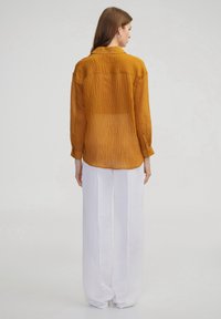 Senfgelbe, gekräuselte Bluse mit langen Ärmeln, Kragen und halbtransparentem Stoff. Kombiniert mit weit geschnittenen weißen Hosen, glatter Textur und minimaler Detailgestaltung.