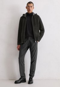 Donkergrijze rits-hoodie met een contrasterend grijs interieur, gedragen over een zwarte coltrui en op maat gemaakte grijze broeken, afgewerkt met zwarte schoenen.