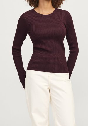 Kobieta w dopasowanym, długim rękawie, prążkowanym bordowym swetrze i luźnych, kremowych spodniach stojąca na jednolitym tle.
