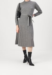 Robe en maille grise à manches longues, jupe plissée, ceinture à la taille et encolure ronde ; associée à des bottines noires et un petit sac à main.