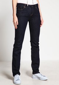 Mörkblå denimjeans med slim fit, med standardfickor och kontrasterande gul sömnad. Kombinerat med vita sneakers.