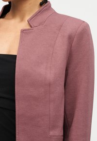 Κοντινό πλάνο ενός ατόμου που φοράει υφασμένο μωβ σακάκι πάνω από μαύρη μπλούζα, δείχνοντας τις λεπτομέρειες γιακά και ώμου.