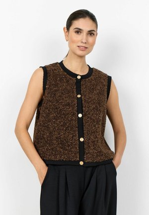 Nő barna, ujjatlan, texturált, gombos mellényben fekete felső és fekete nadrág fölött, kezét zsebből tartva, fehér háttér előtt áll.