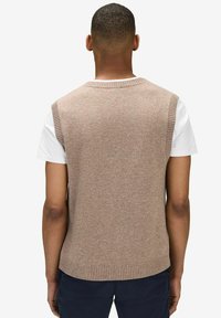 Beige stickad ärmlös tröja med ribbat halsband och fåll, bärs över en vit t-shirt. Slät textur, avslappnad design.