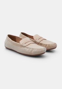 Beige mokasiner av semsket skinn med sydd detaljer, flate såler og en enkel stropp over toppen. Myk tekstur og rund tådesign.
