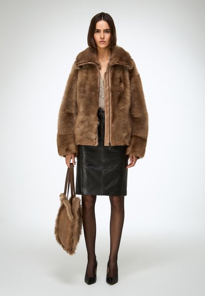Bruine jas van imitatiebont met een rits aan de voorkant, gecombineerd met een zwarte leren kokerrok en zwarte hakken. Model houdt een bijpassende bruine tas vast.