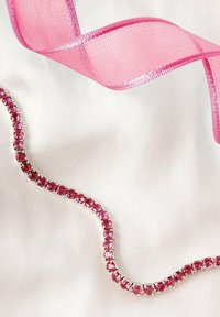 Bordo di strass rosa con una base metallica lucida, forma ondulata, accanto a un nastro rosa trasparente su uno sfondo bianco.