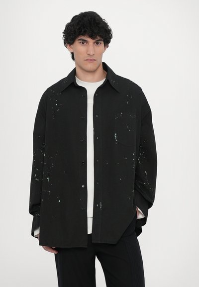Chemise noire à boutons avec des motifs éclaboussés blancs et verts, à manches longues, col et poche poitrine, portée par-dessus un t-shirt blanc.