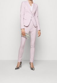 Lila vartota kostyüm, šauniai pritaikytos kelnių, derinamos su balta bluzone. Aksesuarai apima sidabrinę rankinę ir raštuotus aukštakulnius.