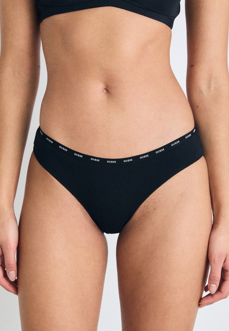 Culottes de bikini noires avec une large taille élastique arborant un logo "GUESS" blanc répété. Tissu lisse avec une couverture minimale.