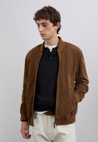 Brown suede bomber jacket with a zipper, ribbed cuffs, and two side pockets. Worn over a black shirt and white trousers.