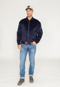 Veste bomber en suede bleu marine avec une fermeture éclair sur le devant, des poignets côtelés et un patch décoratif. Portée avec un jean bleu clair et des bottes grises.