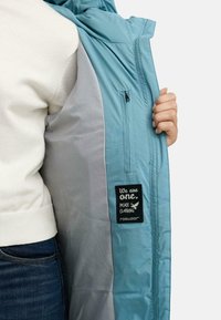 Lyseblå jakke med en glidelås-lomme inni. Inni er det et grått fôr. En svart etikett med hvit tekst leser "Vi er én. Fred Klær."