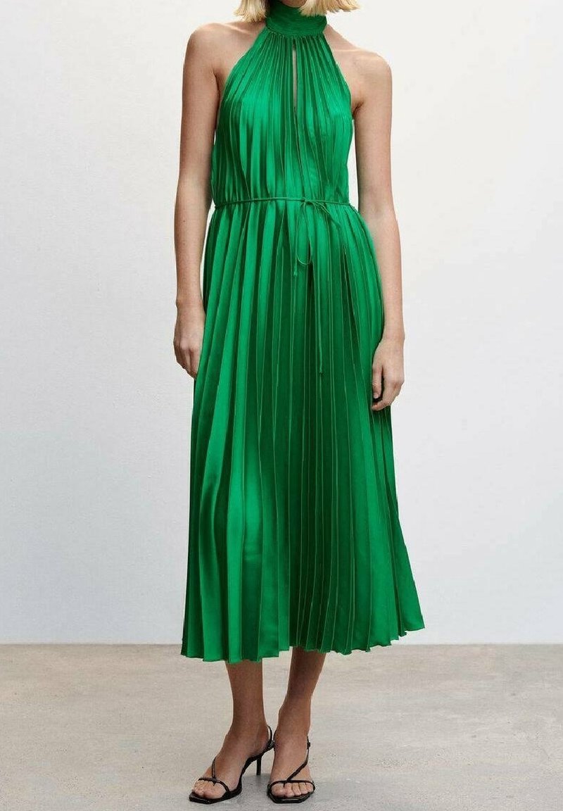 Femme portant une robe midi plissée verte sans manches avec un col montant et une fine ceinture, associée à des talons noirs à brides.