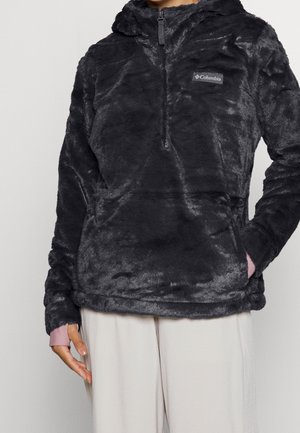 Person iført en sort, blød Columbia hættetrøje med lynlås foran og lyse beige bukser, stående mod en ensfarvet baggrund.