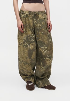 Person iført løstsiddende, vidde bukser med grønt og brunt abstrakt camouflage mønster og brune sko med hvide snørebånd.