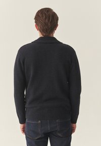 Jūras spēku zils adīts džemperis ar teksturētu šallē kolu un ribotu apakšmalu. Uzvilkts virs tumšiem džinsiem, demonstrējot ikdienišķu piegriezumu un mīkstu audumu.