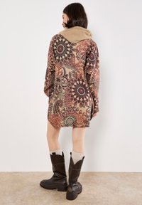 Vestido marrón oversized con estampados y capucha texturizada beige; diseños intrincados en rojo, negro y crema; combinado con botas negras hasta la rodilla.