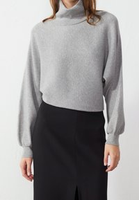 Maglione grigio a collo alto con coste e maniche a palloncino lunghe, abbinato a una gonna a tubino nera con uno spacco laterale. Tessuto morbido, vestibilità comoda.
