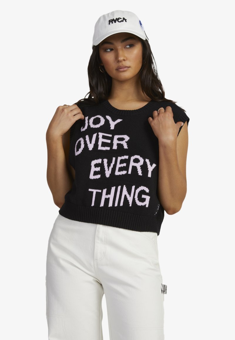 Black crop sweater with pink knitted text "JOY OVER EVERYTHING." Features short sleeves and ribbed hem. Paired with white pants and a cap.