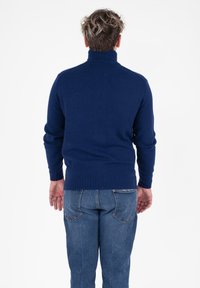 Uomo con capelli ricci corti che indossa un maglione a collo alto blu scuro e jeans blu, in piedi di spalle su uno sfondo bianco.