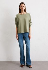 Grüner Pullover mit langen Ärmeln, lässiger Schnitt. Kombiniert mit ausgestellten blauen Jeans und schwarzen Rutschen-Sandalen. Einfaches, minimalistisches Design.