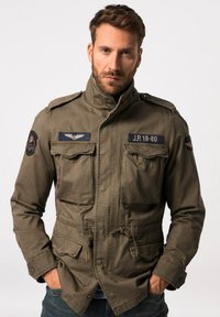 Giacca in stile militare verde oliva, con due tasche sul petto, colletto alto, toppe ricamate e vita regolabile con cordino.