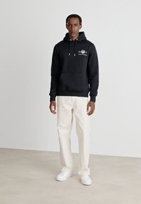 Sort hættetrøje med en frontlomme og hvid grafisk detalje, parret med cremefarvede bukser og hvide sneakers på en neutral baggrund.