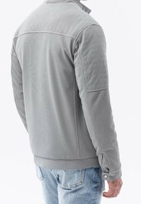 Grijze textuur jasje met een hoge kraag, ritssluiting aan de voorkant en knoopmanchetten. Heeft een getailleerd ontwerp en minimalistische stijl, gecombineerd met lichtblauwe jeans.