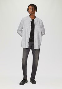 Grijs overhemd met een mandarijnkraag over een zwart shirt met witte stippen, gecombineerd met donkergrijze spijkerbroek en zwarte schoenen, staand tegen een lichte achtergrond.