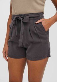 Femme portant un short taille haute gris foncé avec ceinture nouée à l'avant et poches, accompagné d'un haut beige sans manches.