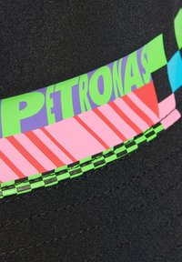 Tela negra con una franja colorida que presenta la palabra "PETRONAS" en verde y morado, rayas diagonales en rosa y rojo, y un patrón de cuadros verdes y negros.