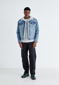 Veste en denim bleu clair avec un col en shearling beige, pull gris, pantalon noir et chaussures vertes. Le modèle est posé contre un fond uni.