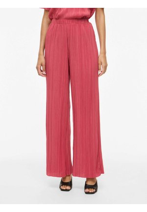 Femme portant un pantalon rose plissé taille haute, à jambes larges, associé à des sandales à talons noires ouvertes à l'avant, sur un fond uni.