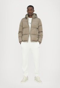 Puffer jacket in light brown, featuring a zip front, two side pockets, and a hood. Worn with white pants and light-colored sneakers.