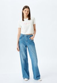 Witte cropped t-shirt met roze strikafbeeldingen, gecombineerd met lichtblauwe denim jeans met wijde pijpen. Model staat met één hand in de zak van de jeans.