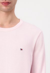 Gros plan d'une personne portant un pull rose clair avec un petit logo rectangulaire rouge, blanc et bleu sur la poitrine.