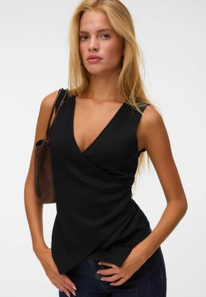 Blonde woman with long hair wearing a sleeveless black V-neck top and dark pants, carrying a brown shoulder bag.