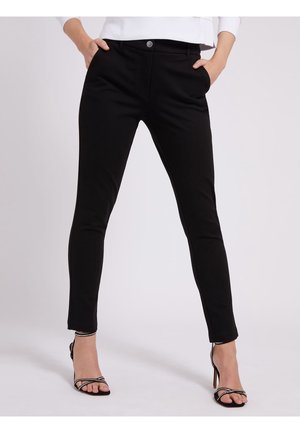 Frau trägt schmale schwarze Hose und schwarze Sandalen mit Absatz und Strassriemen, steht mit den Händen in den Taschen vor weißem Hintergrund.