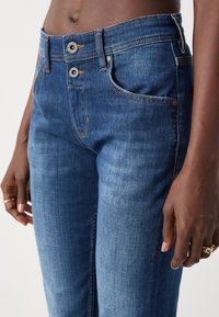 Vaqueros de denim azul de cintura alta, con cierre de dos botones y costuras en contraste. Incluyen bolsillos laterales y un ajuste entallado. Textura suave.