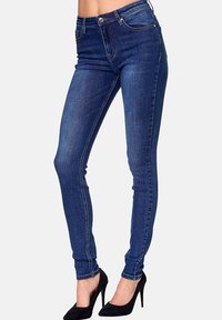 Jeans ajustados en denim azul oscuro, con bolsillos frontales y traseros, costuras en contraste y un diseño ajustado. Combinados con tacones altos negros.