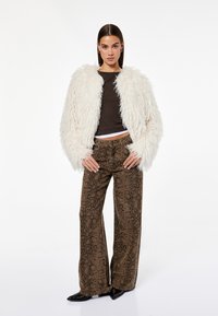 Kurtka z sztucznego futra w kolorze kremowym, brązowa bluzka, wzorzyste szerokie spodnie w brązowe i czarne wzory oraz czarne buty na szpilkach.