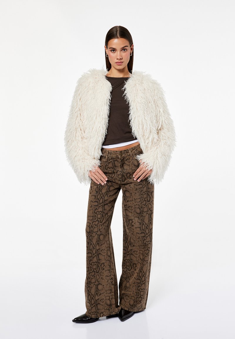 Kurtka z sztucznego futra w kolorze kremowym, brązowa bluzka, wzorzyste szerokie spodnie w brązowe i czarne wzory oraz czarne buty na szpilkach.