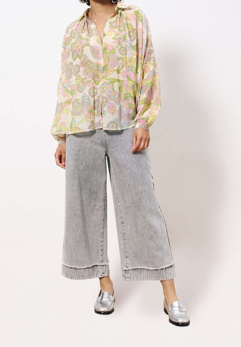 Blouse à motif floral aux couleurs pastel avec manches longues, portée avec un jean gris à jambes larges légèrement effiloché et des mocassins argentés.