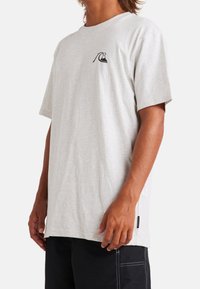 Camiseta de algodón gris claro con mangas cortas, que presenta un pequeño logo negro de montaña y ola en el pecho y una etiqueta lateral.