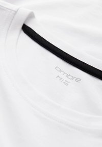 Witte katoenen T-shirt met een zwarte geribbelde halslijn. Het label toont "Ombre M 48" in grijze letters op de stof. Soepele textuur, casual ontwerp.
